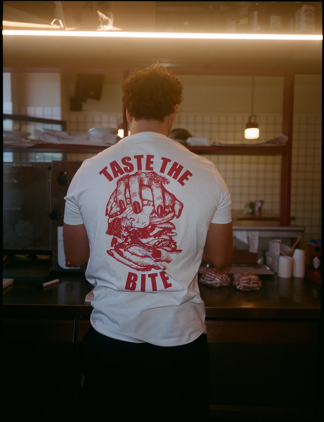 Behind the counter: a staff T-shirt printed “Taste the bite”, cream tiles, red steel shelving and burgers wrapped in striped paper under warm light.