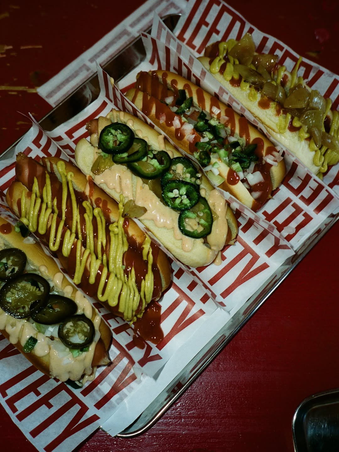 Four hot dogs dressed four ways — jalapeños, mustard, ketchup, grilled onion — lined up on LEFTY-printed paper.