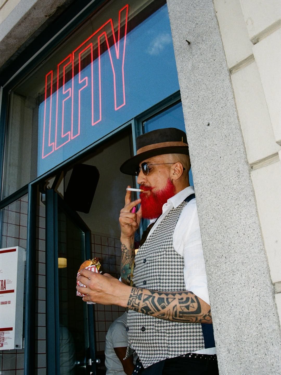 A regular in a hat and sunglasses, beard dyed red, bites into a wrapped burger beneath the red neon LEFTY sign in the window.